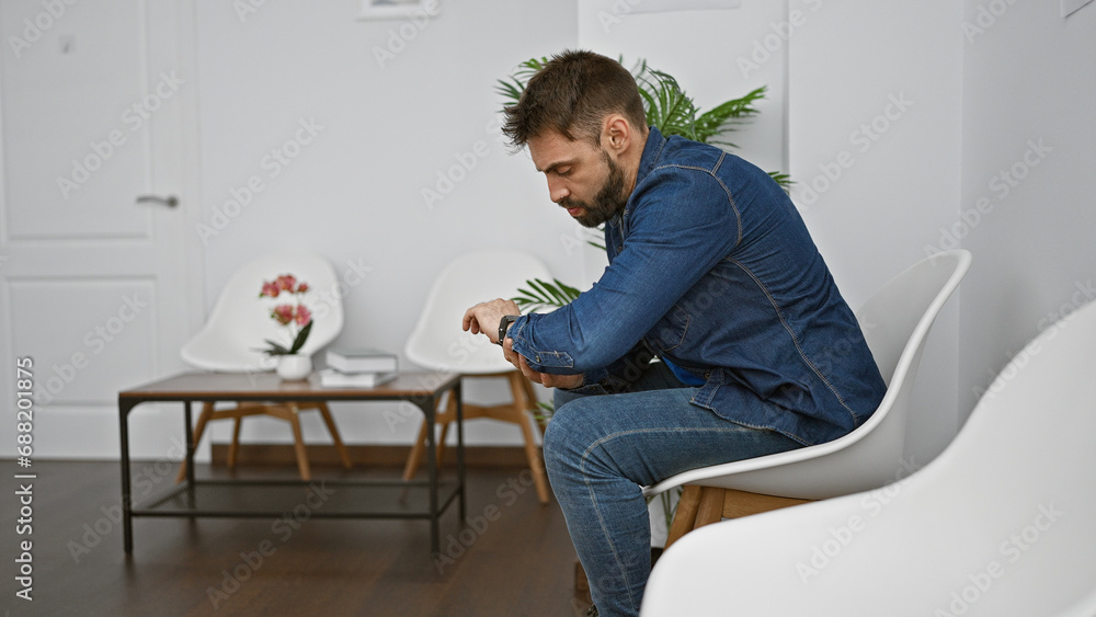 Casual yet serious, a young hispanic man sits relaxed in a waiting room chair, sporting a handsome beard. his intense expression, as he concentrates on a watch, hints at a problem. is it a countdown?