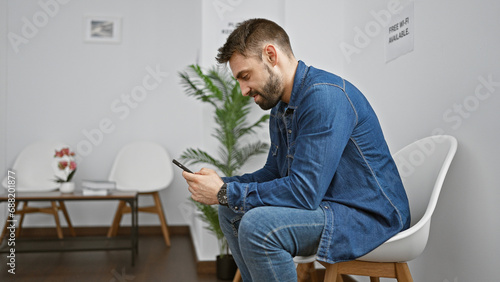 Handsome young hispanic male chilling in the waiting room, immersed in his smartphone screen, casually typing messages. a portrait of relaxation and concentration amidst the indoor background.