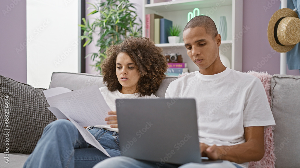 Beautiful couple in love, sitting together on their sofa at home, seriously concentrated on reading a bill on their laptop, embracing relaxed lifestyle indoors. Beautiful couple in love, sitting together on their sofa at home, seriously concentrated on reading a bill on their laptop, embracing relaxed lifestyle indoors.