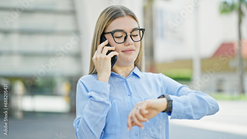 Young blonde woman business worker talking on smartphone looking watch at street