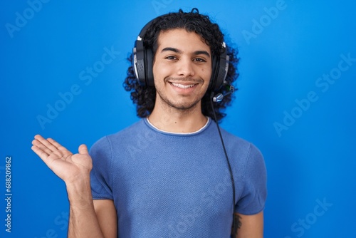 Hispanic man with curly hair listening to music using headphones smiling cheerful presenting and pointing with palm of hand looking at the camera.