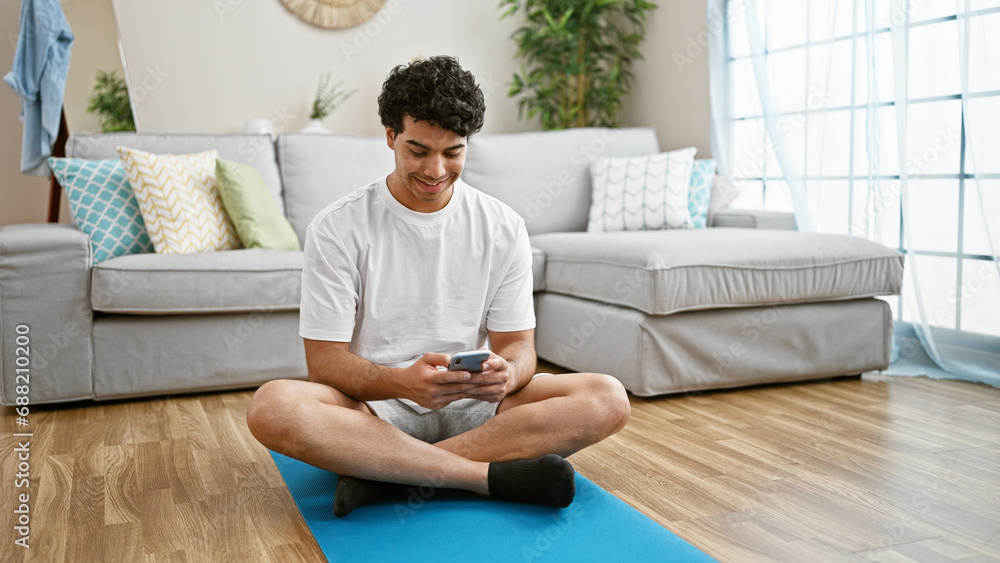 Young latin man using smartphone sitting on yoga mat at home