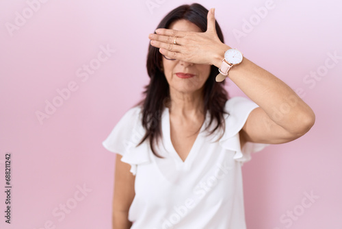 Middle age hispanic woman wearing casual white t shirt and glasses covering eyes with hand, looking serious and sad. sightless, hiding and rejection concept