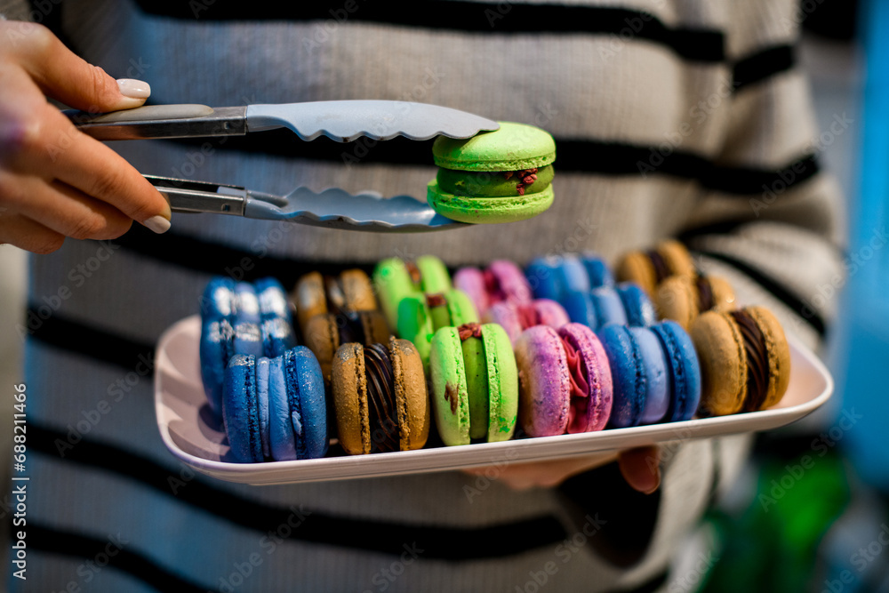 Naklejka premium Female hands hold a set of colorful macaroons and in the other hand one macaroon with kitchen tongs
