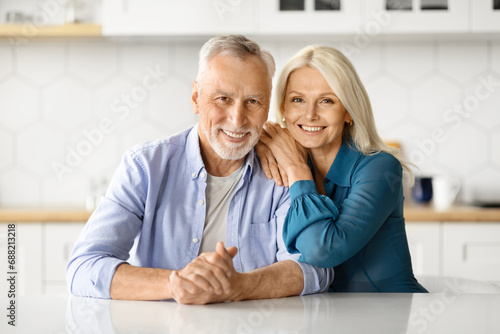 Portrait Of Happy Senior Spouses Posing At Table In Kitchen 