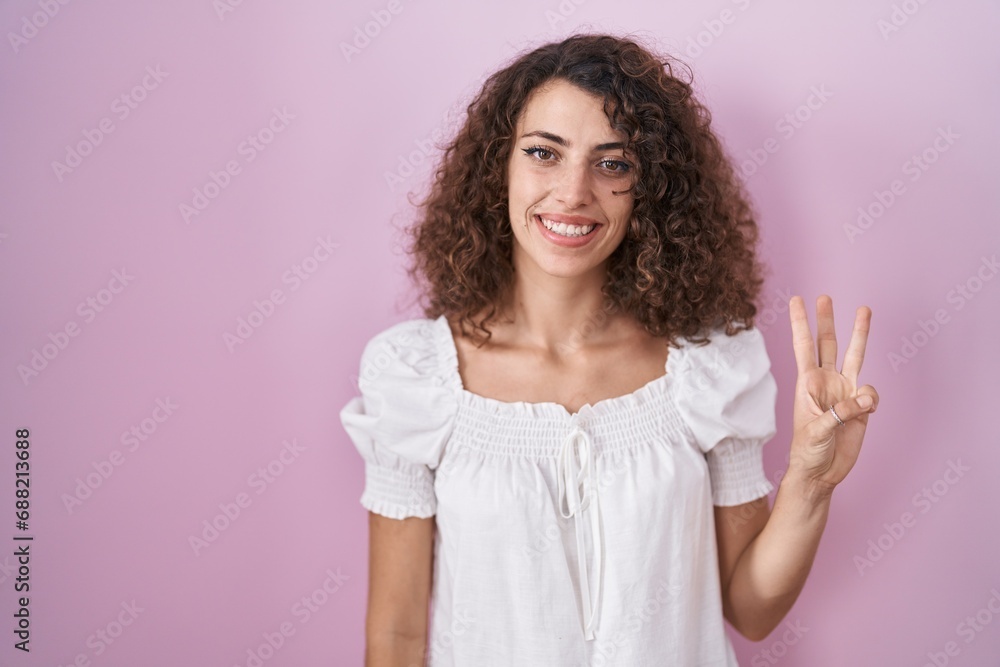 Hispanic woman with curly hair standing over pink background showing and pointing up with fingers number three while smiling confident and happy. Hispanic woman with curly hair standing over pink background showing and pointing up with fingers number three while smiling confident and happy.