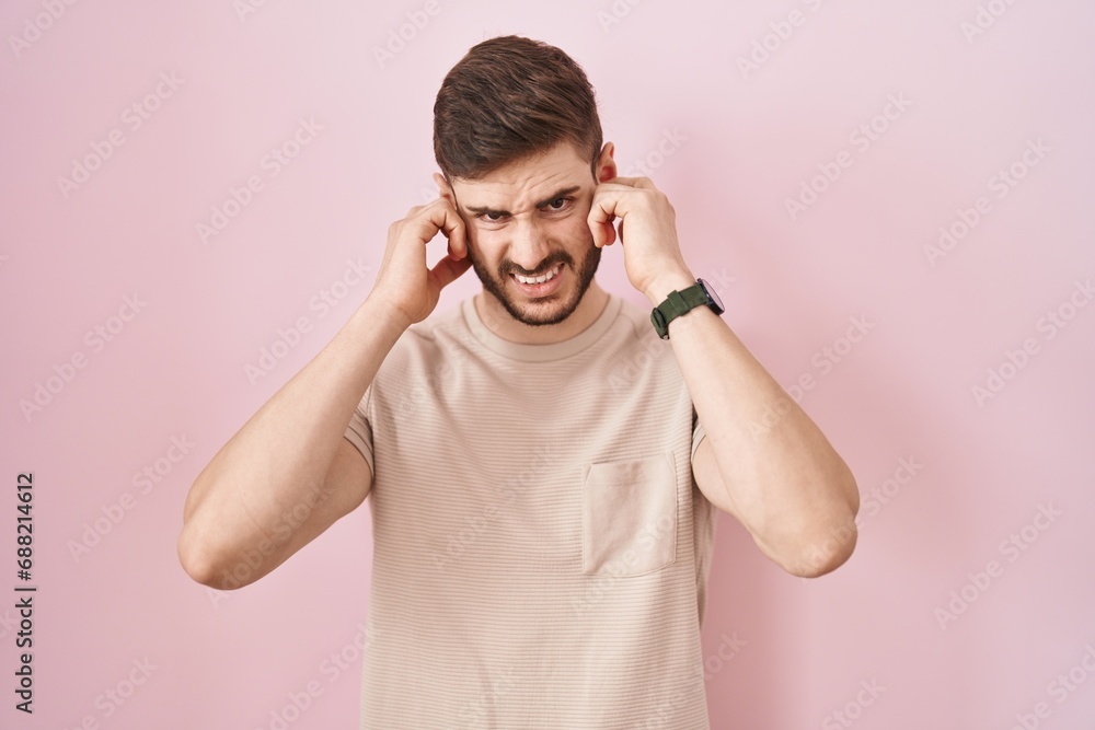 Hispanic man with beard standing over pink background covering ears with fingers with annoyed expression for the noise of loud music. deaf concept. Hispanic man with beard standing over pink background covering ears with fingers with annoyed expression for the noise of loud music. deaf concept.