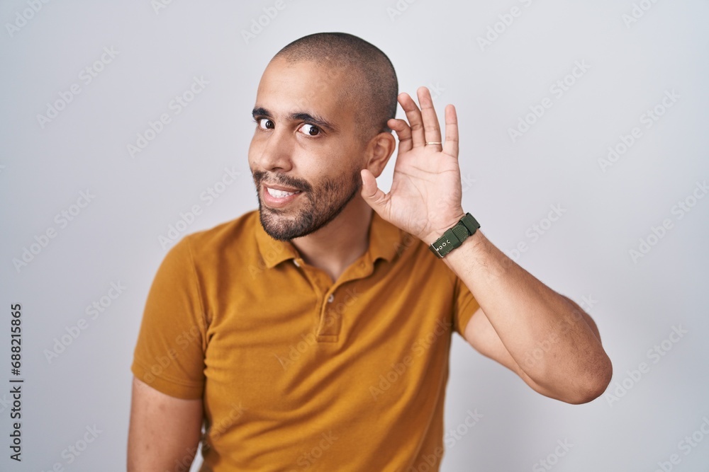 Hispanic man with beard standing over white background smiling with hand over ear listening an hearing to rumor or gossip. deafness concept. Hispanic man with beard standing over white background smiling with hand over ear listening an hearing to rumor or gossip. deafness concept.