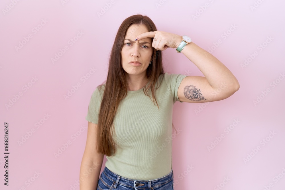 Beautiful brunette woman standing over pink background pointing unhappy to pimple on forehead, ugly infection of blackhead. acne and skin problem