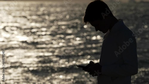 silhouette of lonely guy by the sea at sunset scrolling the phone keypad photo