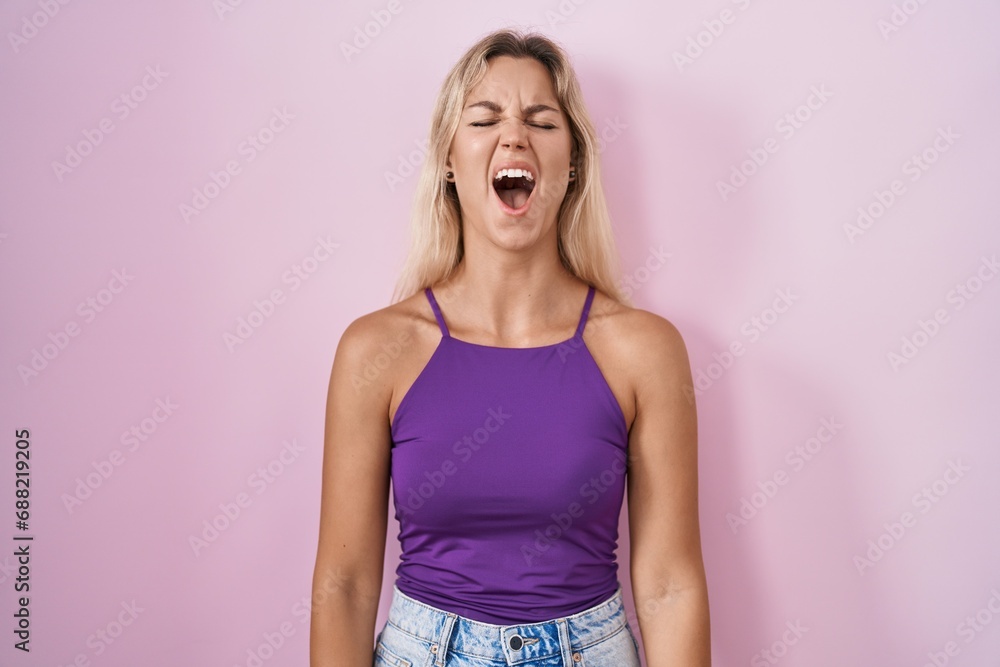 Young blonde woman standing over pink background angry and mad screaming frustrated and furious, shouting with anger. rage and aggressive concept. Young blonde woman standing over pink background angry and mad screaming frustrated and furious, shouting with anger. rage and aggressive concept.