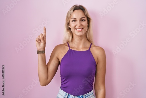 Young blonde woman standing over pink background showing and pointing up with finger number one while smiling confident and happy. © Krakenimages.com