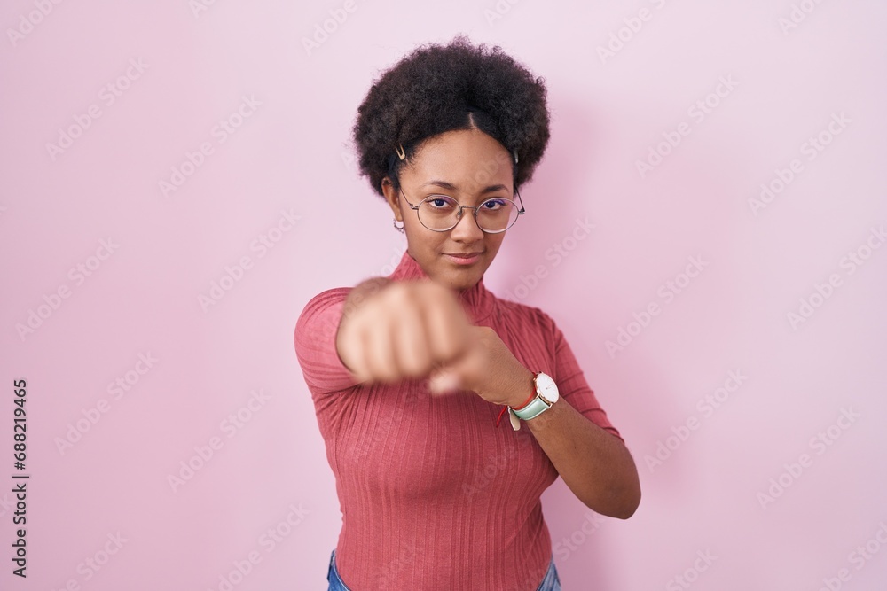 Beautiful african woman with curly hair standing over pink background punching fist to fight, aggressive and angry attack, threat and violence
