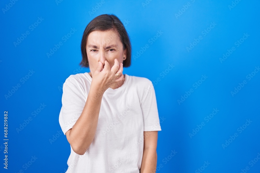Middle age hispanic woman standing over blue background smelling something stinky and disgusting, intolerable smell, holding breath with fingers on nose. bad smell Middle age hispanic woman standing over blue background smelling something stinky and disgusting, intolerable smell, holding breath with fingers on nose. bad smell