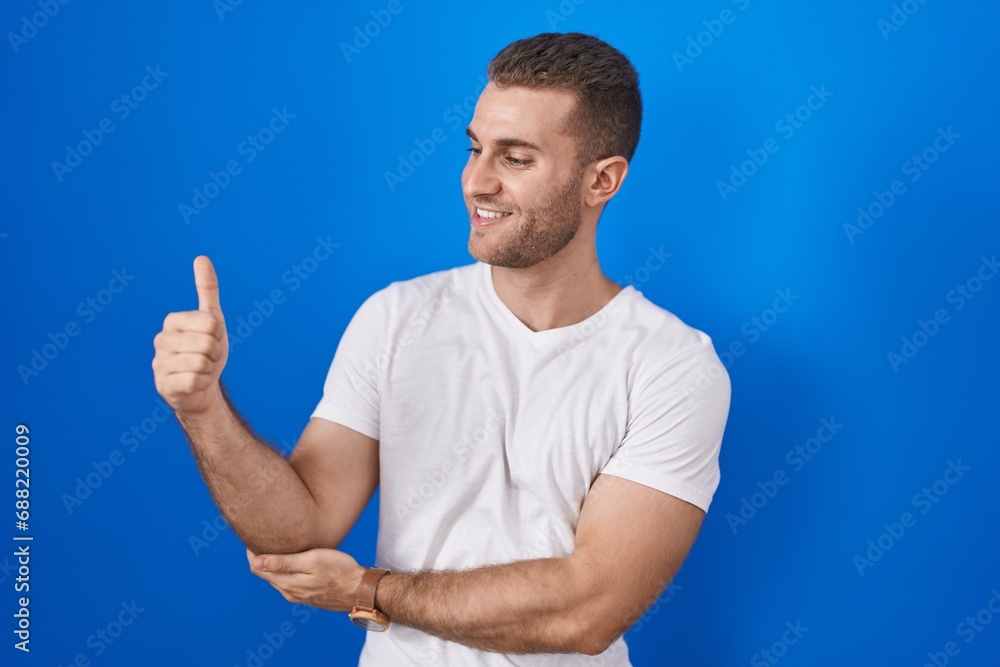 Fototapeta premium Young caucasian man standing over blue background looking proud, smiling doing thumbs up gesture to the side