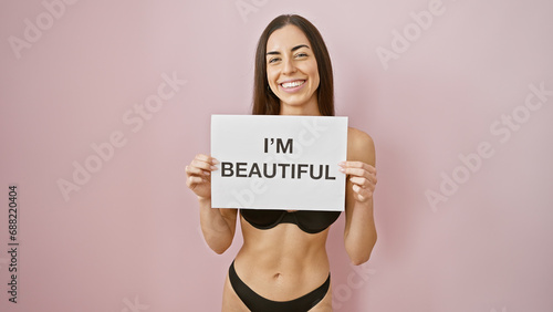 Sassy morning energy! young, confident, hispanic woman, sexily standing in lingerie, holding an 'i'm beautiful' banner, flaunting her hairstyle over an isolated pink background.