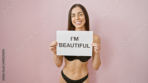 Sassy morning energy! young, confident, hispanic woman, sexily standing in lingerie, holding an 'i'm beautiful' banner, flaunting her hairstyle over an isolated pink background. photo