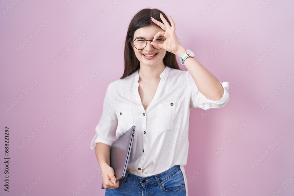 Young caucasian woman holding laptop doing ok gesture with hand smiling, eye looking through fingers with happy face. Young caucasian woman holding laptop doing ok gesture with hand smiling, eye looking through fingers with happy face.
