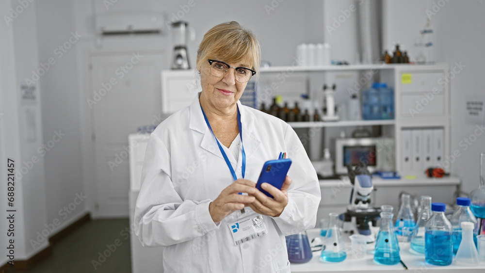 Blonde middle age woman scientist, all smiles as she efficiently uses her smartphone amidst work in the lab. Blonde middle age woman scientist, all smiles as she efficiently uses her smartphone amidst work in the lab.
