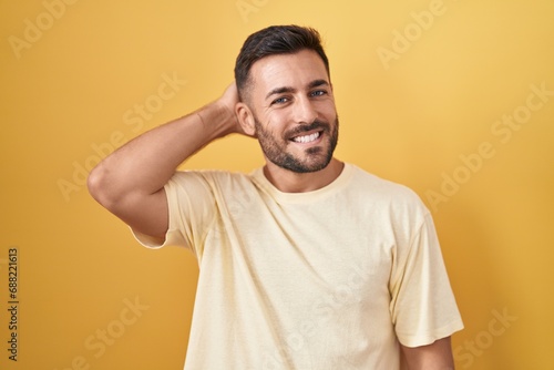 Handsome hispanic man standing over yellow background smiling confident touching hair with hand up gesture, posing attractive and fashionable