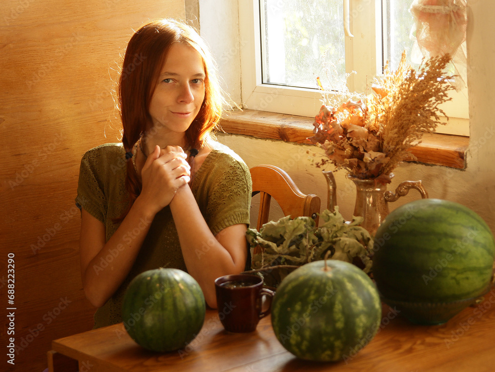 Fototapeta premium Selfie portrait of fair-haired woman in ethnic natural clothes in a country house, in the rays of the sun. What is sitting at the table on which there are large green watermelons.