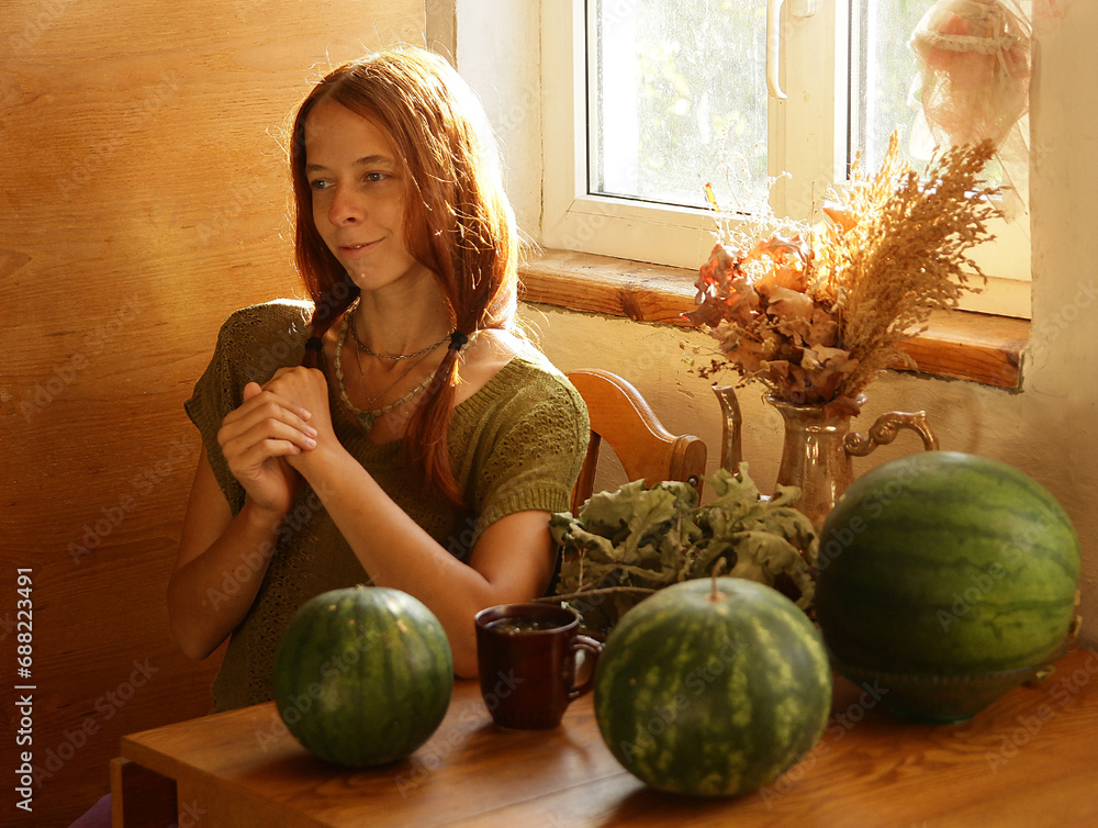 Fototapeta premium Selfie portrait of fair-haired woman in ethnic natural clothes in a country house, in the rays of the sun. What is sitting at the table on which there are large green watermelons.