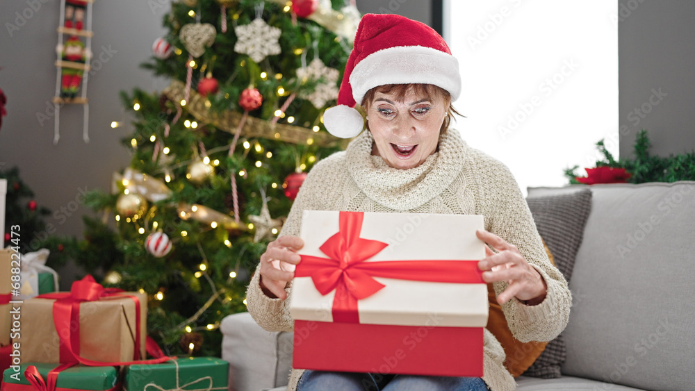 Mature hispanic woman wearing christmas hat unpacking gift with surprised face at home
