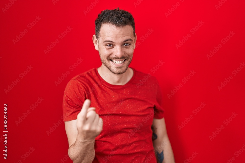 Young hispanic man standing over red background beckoning come here gesture with hand inviting welcoming happy and smiling Young hispanic man standing over red background beckoning come here gesture with hand inviting welcoming happy and smiling