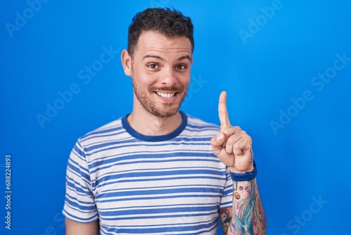 Young hispanic man standing over blue background showing and pointing up with finger number one while smiling confident and happy. © Krakenimages.com