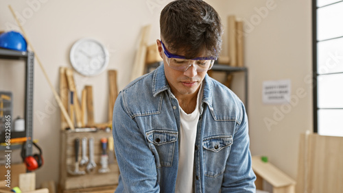 Handsome, young hispanic man standing tall in carpentry work studio â€“ a portrait of dedication and seriousness. this glasses-clad, professional carpenter is a master in the realm of woodworking