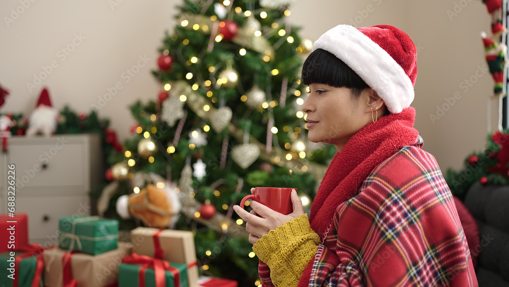 Young chinese woman drinking coffee sitting on sofa by christmas tree at home