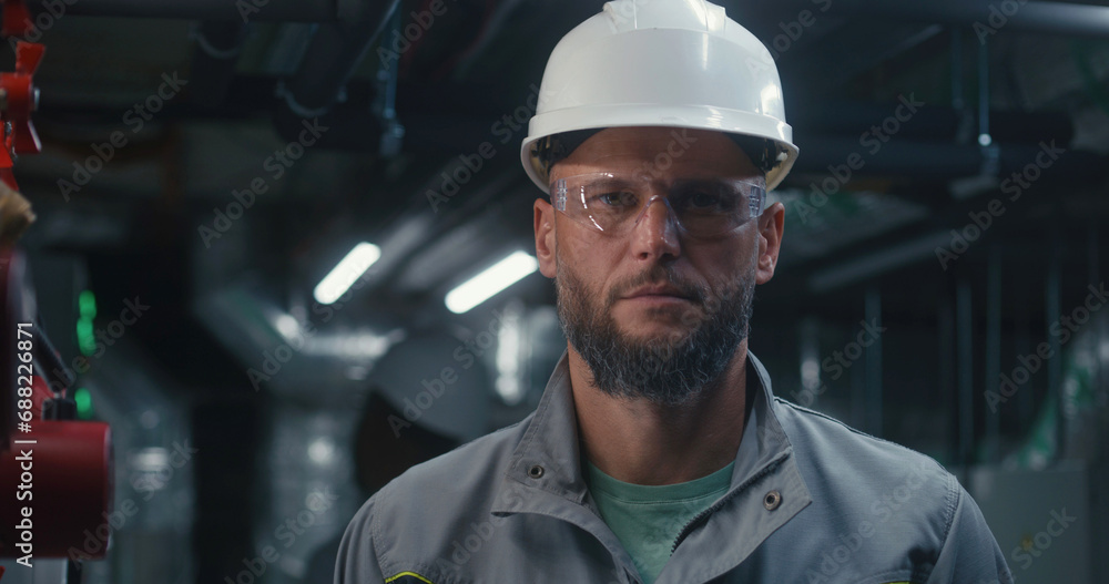 Professional Caucasian engineer stands and looks at camera. Industrial specialist or worker in safety uniform, goggles and hard hat works on modern manufacturing factory, plant or energy facility. Professional Caucasian engineer stands and looks at camera. Industrial specialist or worker in safety uniform, goggles and hard hat works on modern manufacturing factory, plant or energy facility.