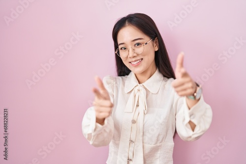 Young chinese woman standing over pink background pointing fingers to camera with happy and funny face. good energy and vibes.