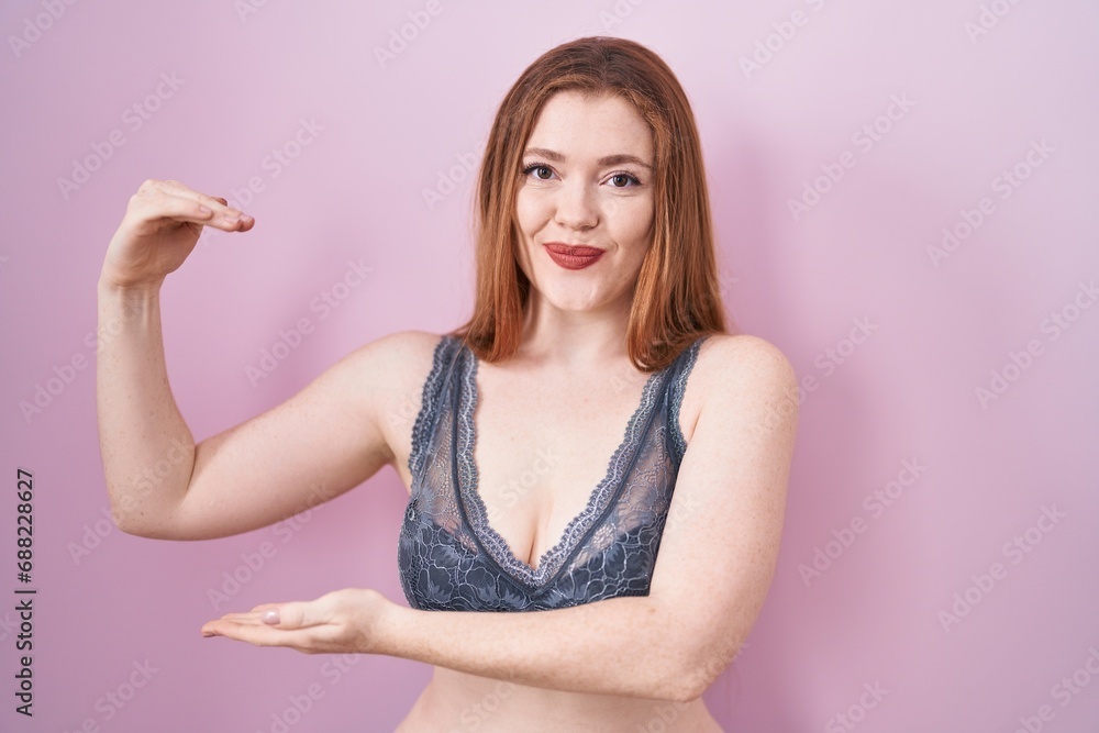 Redhead woman wearing lingerie over pink background gesturing with hands showing big and large size sign, measure symbol. smiling looking at the camera. measuring concept. Redhead woman wearing lingerie over pink background gesturing with hands showing big and large size sign, measure symbol. smiling looking at the camera. measuring concept.