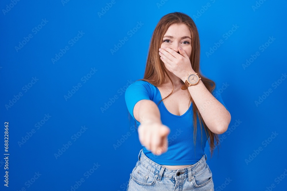 Redhead woman standing over blue background laughing at you, pointing finger to the camera with hand over mouth, shame expression Redhead woman standing over blue background laughing at you, pointing finger to the camera with hand over mouth, shame expression