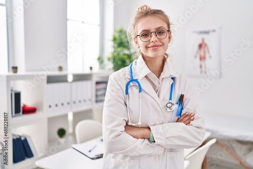Young blonde woman doctor smiling confident standing with arms crossed gesture at clinic