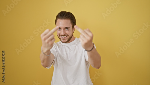 Young hispanic man smiling confident doing middle finger gesture over isolated yellow background
