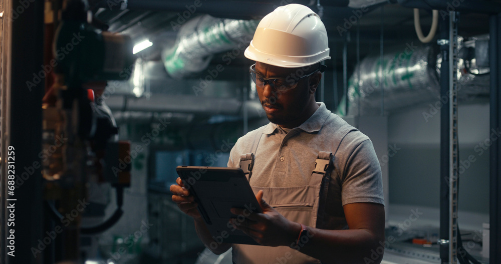 Male technician worker wearing safety uniform and hard hat works using tablet computer. African American inspector checks pipeline system on modern factory, plant or industrial energy facility. Male technician worker wearing safety uniform and hard hat works using tablet computer. African American inspector checks pipeline system on modern factory, plant or industrial energy facility.