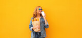 Portrait of happy smiling young woman holding grocery shopping paper bag with long white bread baguette and cup of coffee on orange background