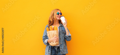 Portrait of happy smiling young woman holding grocery shopping paper bag with long white bread baguette and cup of coffee on orange background