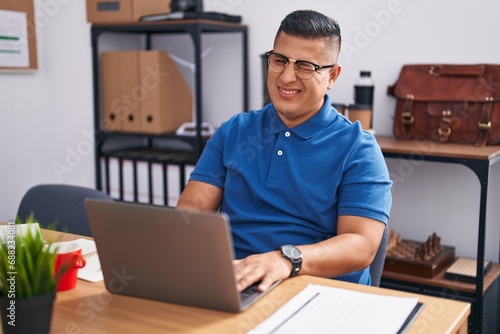 Young hispanic man working at the office with laptop winking looking at the camera with sexy expression, cheerful and happy face.