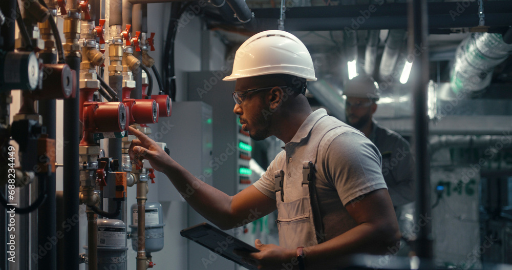 Male technician worker wearing safety uniform and hard hat works using tablet computer. African American inspector checks pipeline system on modern factory, plant or industrial energy facility. - obrazy, fototapety, plakaty 