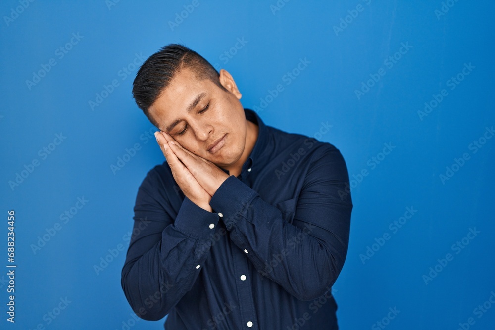 Hispanic young man standing over blue background sleeping tired dreaming and posing with hands together while smiling with closed eyes. Hispanic young man standing over blue background sleeping tired dreaming and posing with hands together while smiling with closed eyes.