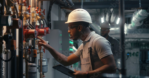 Male technician worker wearing safety uniform and hard hat works using tablet computer. African American inspector checks pipeline system on modern factory, plant or industrial energy facility. photo