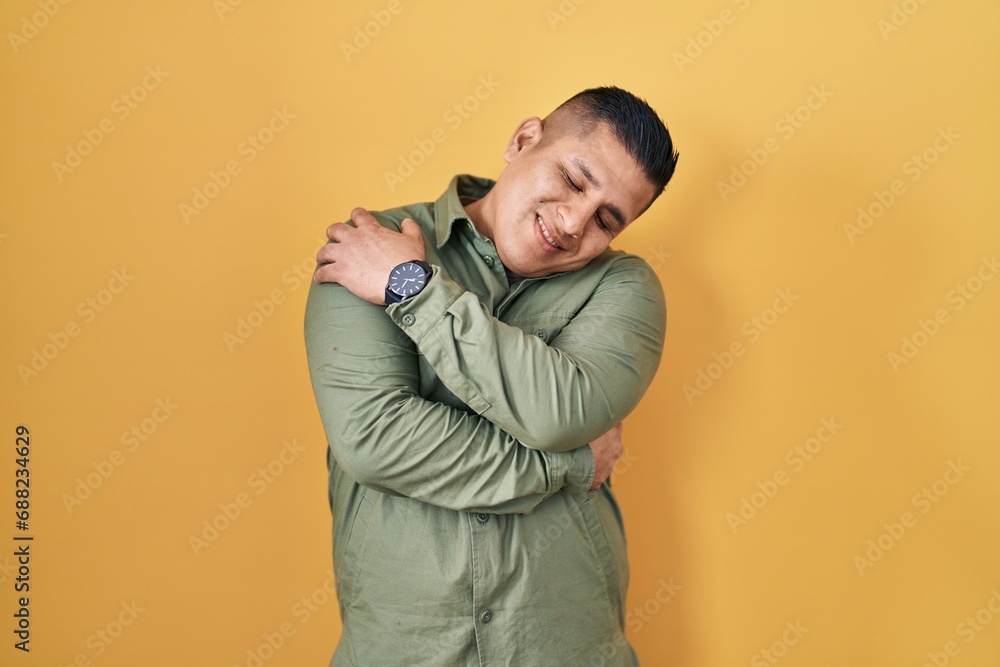 Hispanic young man standing over yellow background hugging oneself happy and positive, smiling confident. self love and self care Hispanic young man standing over yellow background hugging oneself happy and positive, smiling confident. self love and self care