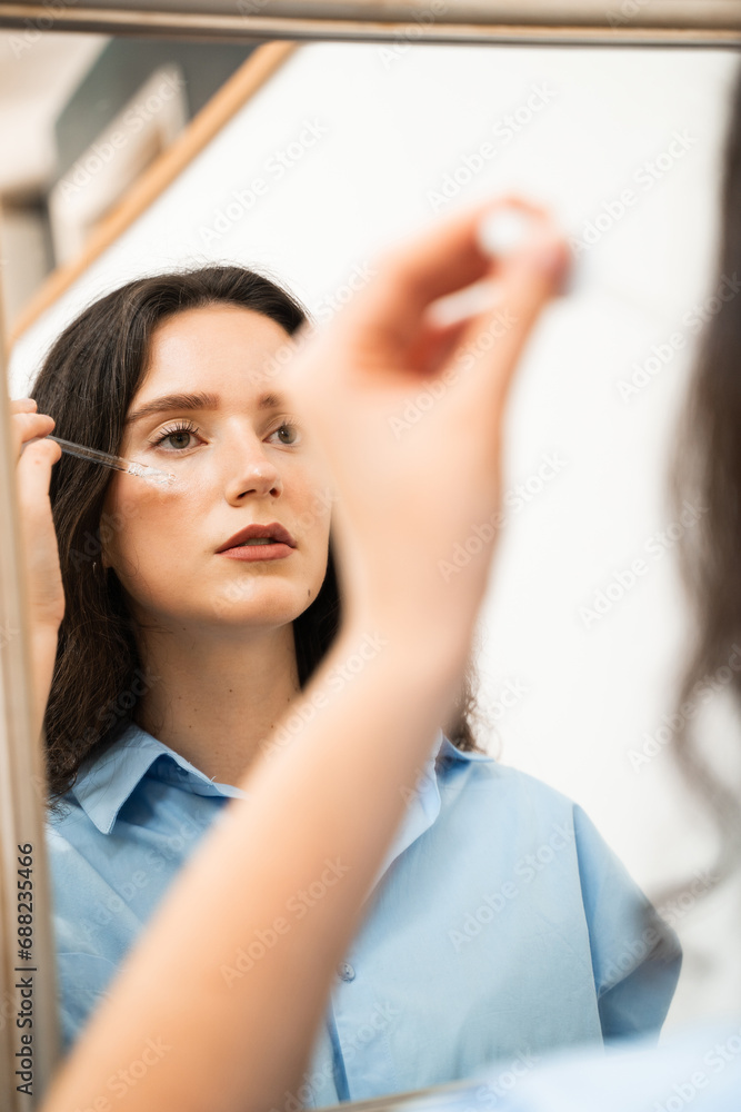 Fototapeta premium Girl with hyaluronic acid or serum pipette in hands is looking in the mirror. Young woman is applying moisturizing serum on skin face.