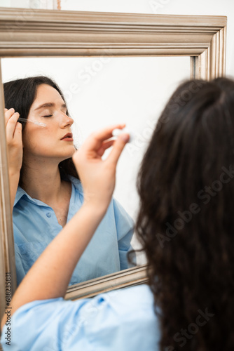 Young woman is applying moisturizing serum on skin face is looking in the mirror. Girl with hyaluronic acid or serum pipette in hands close-up.