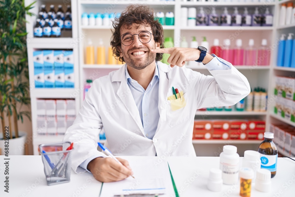 Hispanic young man working at pharmacy drugstore smiling cheerful showing and pointing with fingers teeth and mouth. dental health concept.