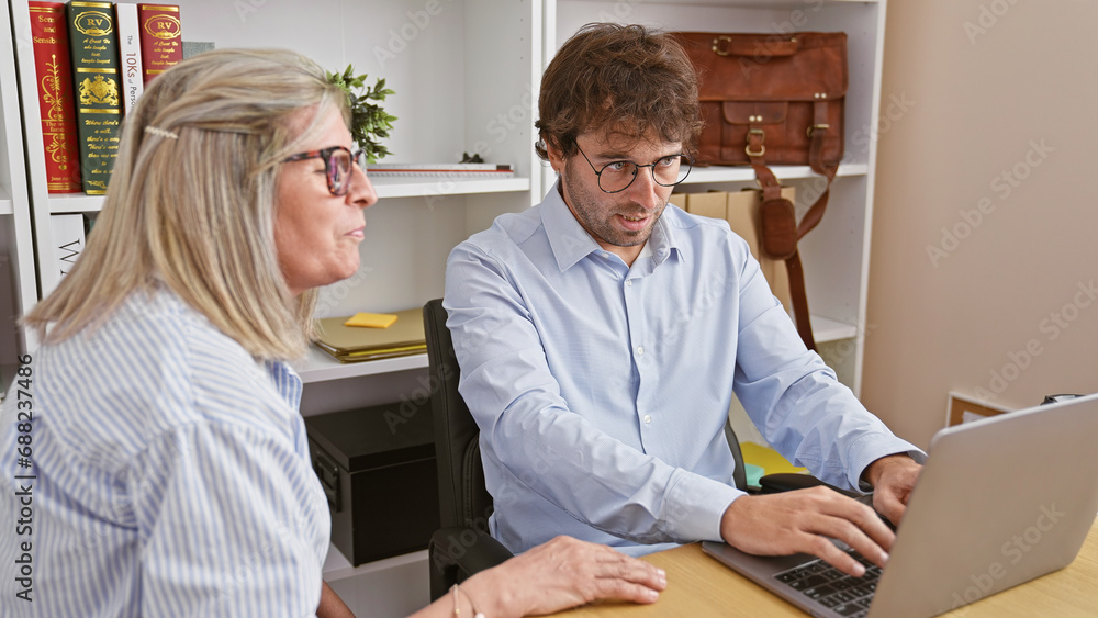 Two hardworking office workers, a man and a woman, together intensely working on a laptop, projecting the image of an unstoppable team. Two hardworking office workers, a man and a woman, together intensely working on a laptop, projecting the image of an unstoppable team.