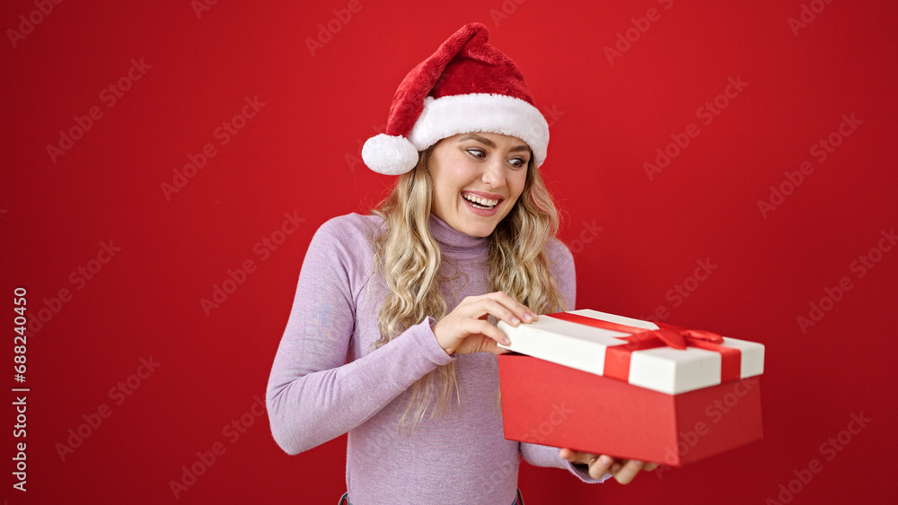 Young blonde woman wearing christmas hat unpacking gift over isolated red background Young blonde woman wearing christmas hat unpacking gift over isolated red background